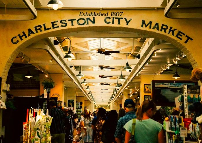 A bustling indoor market with shoppers all around. An arch above reads, 'Established 1807 Charleston City Market'