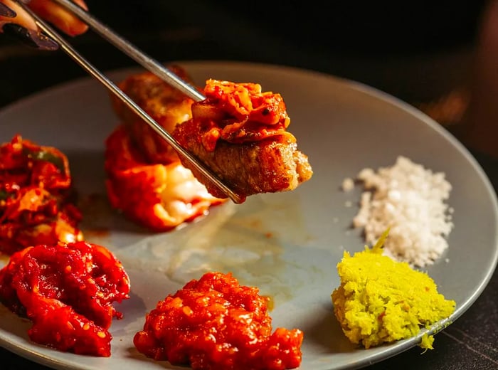 A pair of metal chopsticks holds a grilled meat slice topped with kimchi, positioned before a plate of assorted banchan.