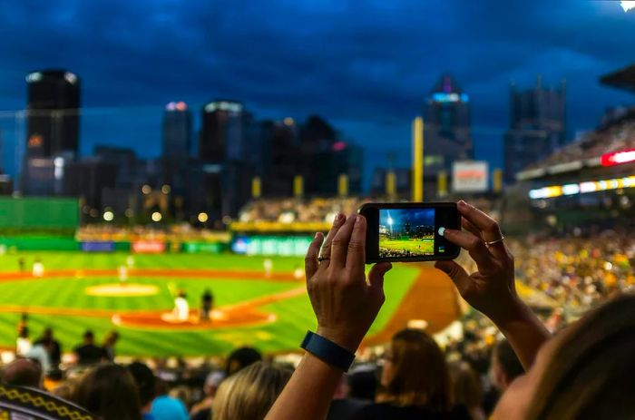 A woman captures a photo of a Pirates baseball game at PNC Park, framed by the dazzling Pittsburgh skyline at night.