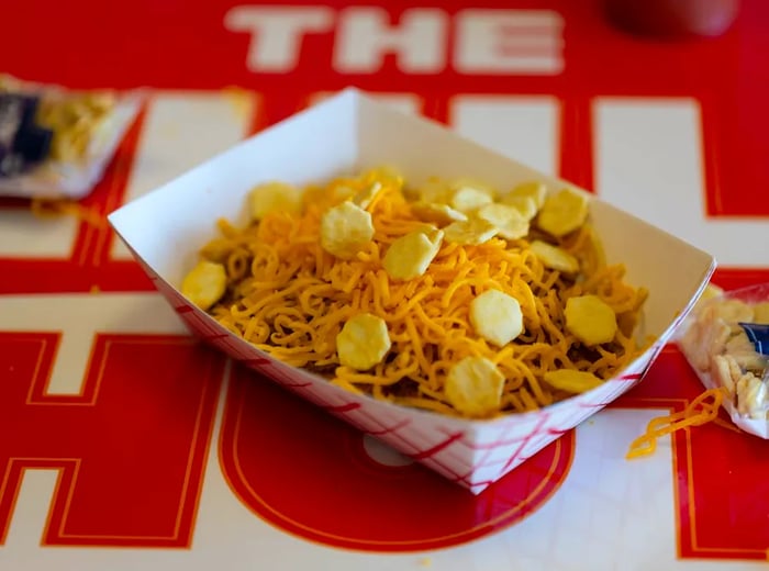 A small cardboard boat filled with cheese and crackers, resting atop a hidden layer of spaghetti and chili.