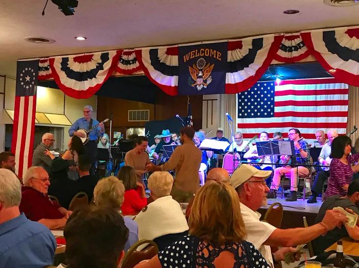 A stage adorned with the red, white, and blue of the U.S. flag is filled with seated musicians. In front of the stage, two individuals are smiling and dancing.