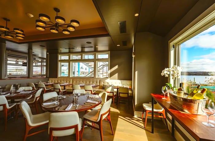 A sunlit dining area showcasing ocean views, large tables set for an evening meal, and contemporary chandeliers overhead.