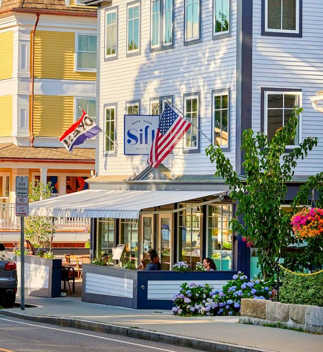 A charming bakery facade on a sunny street, where diners enjoy meals at outdoor tables under an awning, surrounded by several flags.