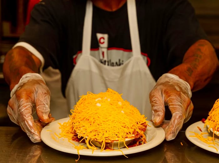 A cook wearing a branded Camp Washington shirt serves spaghetti topped with cheese and crackers across the pass.