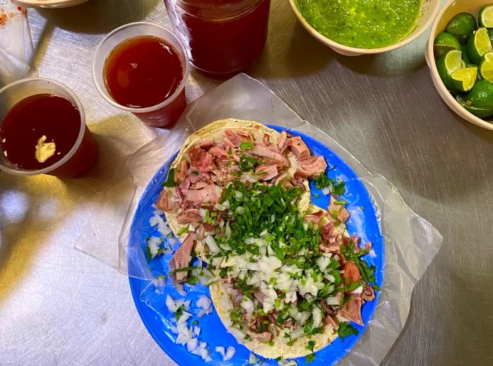 From above, two tacos generously topped with cilantro and onions, resting on a blue plate next to salsas, lime wedges, and various sauces.