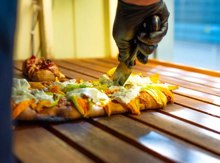 An employee wearing gloves carefully slices a pizza on a wooden counter beside a sunlit window.