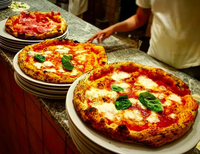 A chef stands beside an array of whole pizzas, prominently featuring two margheritas.