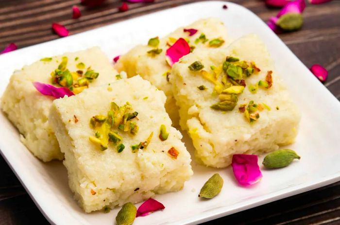 Indian Mithai, particularly hot Kalakand Burfi—also known as Alwar Mawa Qalaqand, Mishri Mava, or Khoa Milk Cake—is made from solidified milk and topped with crushed pistachios and cardamom.