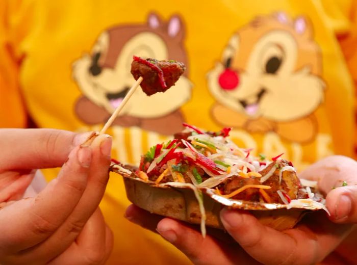 A hand lifts a piece of potato on a skewer from a bowl of aloo chaat.