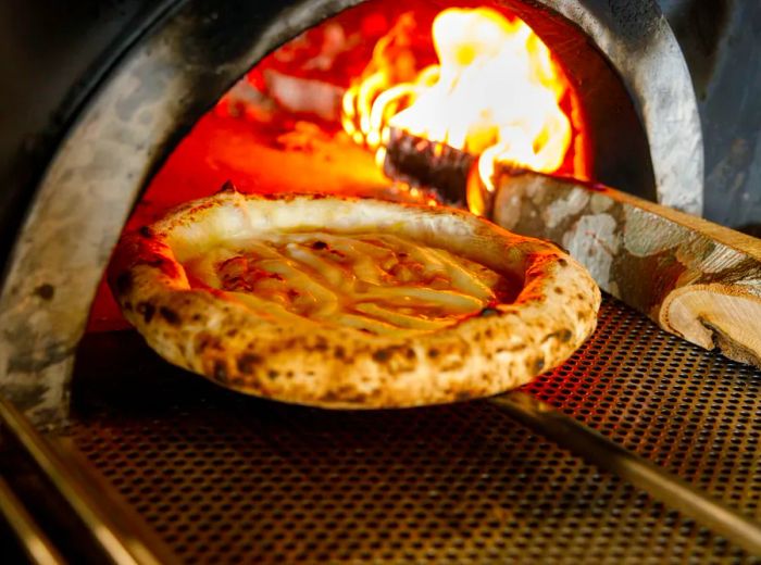 A pizza rests on a wooden paddle at the entrance of a blazing oven.