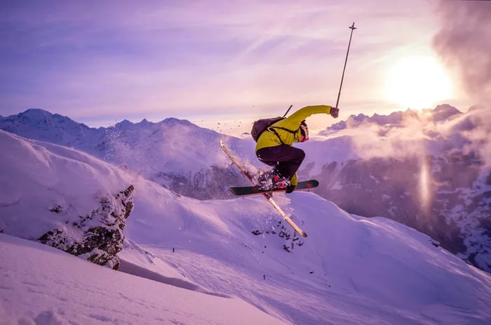 A skier soars through the air on the slopes overlooking Verbier in the Swiss Alps, where fresh snow blankets the mountains and the sun sets behind the peaks.