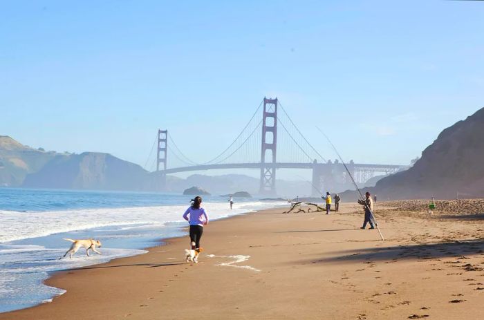 Individuals jogging and fishing on Baker Beach near the Golden Gate Bridge.