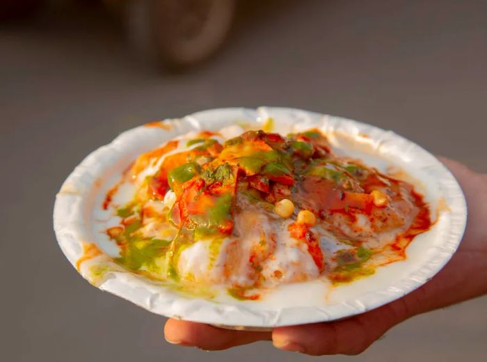 A hand holds a paper bowl filled with papdi chaat adorned with vibrant chutneys.