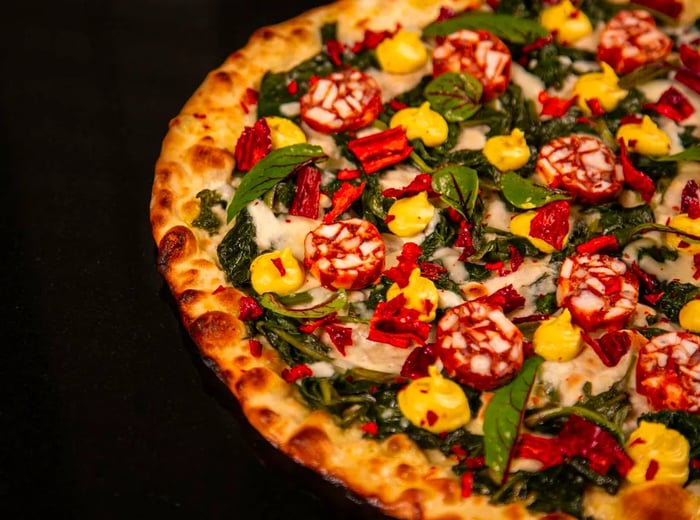 A partial view of a pizza on a black background, topped with chorizo, peppers, and sorrel.