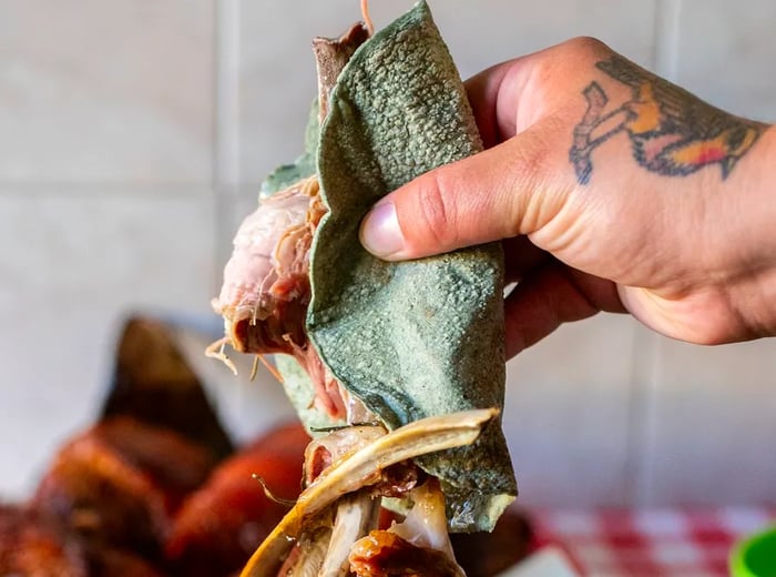 A hand reaches for meat from a large piece of roasted barbacoa, wrapped in a blue tortilla.