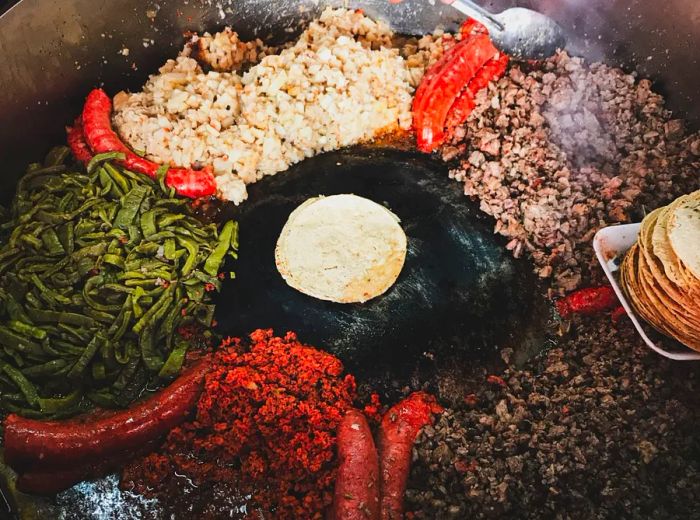 A taquero stands over a large comal, where various meats and vegetables sizzle away, with a single tortilla toasting at the center.