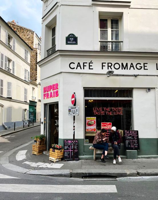 Two customers relax on a bench outside a corner grocery, surrounded by piles of fresh produce and charming chalkboard signs.