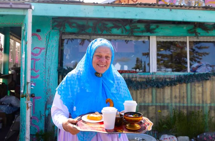 A cheerful older woman in a blue headscarf carries a tray filled with tea.