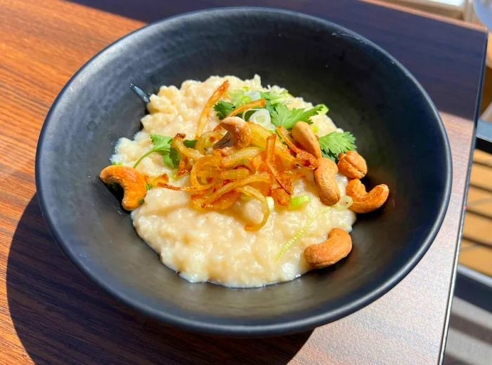 A bowl of congee topped with cashews, shallots, and cilantro on a table.