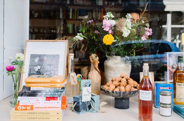 A window display seen from the outside features a stack of books, a bowl of chestnuts, various bottles and cans, along with a vase of flowers.