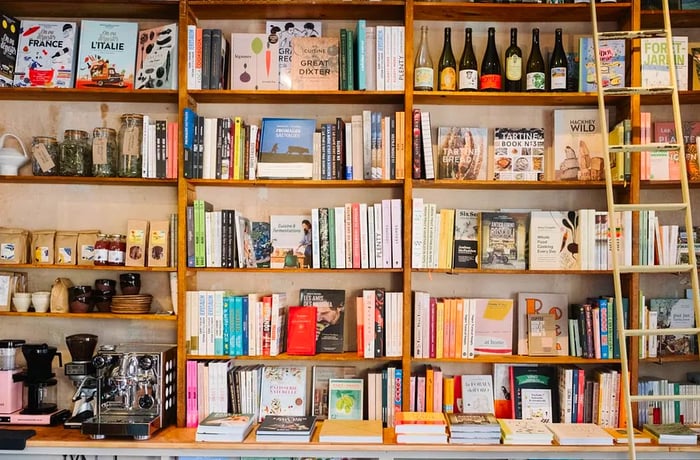 Tall bookshelves are lined with books, wine bottles, an espresso machine, and bags of coffee, with a ladder positioned at one end.