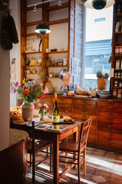 A candle flickers in an empty wine bottle on a table next to a pot of flowers and food-related books. The sunlit interior reveals shelves filled with various items and dark wood drawers beyond.