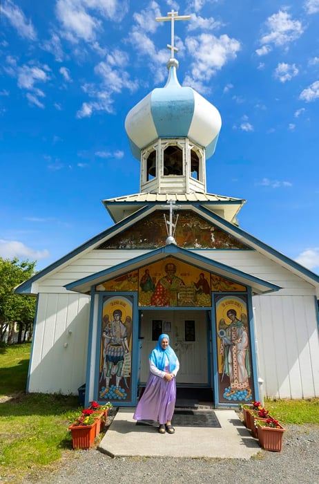 A woman wearing a blue headscarf stands before a small white church, topped with a blue and white striped dome, adorned with frescos around its doorways.