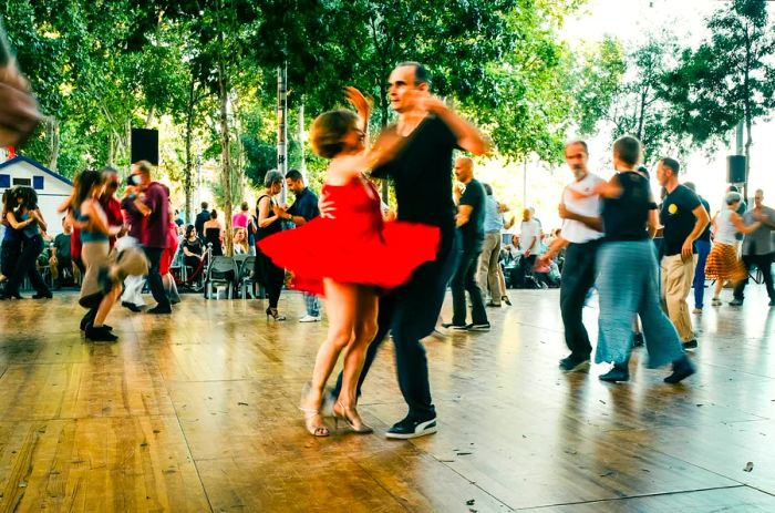 A couple dances energetically in a public square during the Tangopostale festival in Toulouse, France, featuring a woman in a red dress.