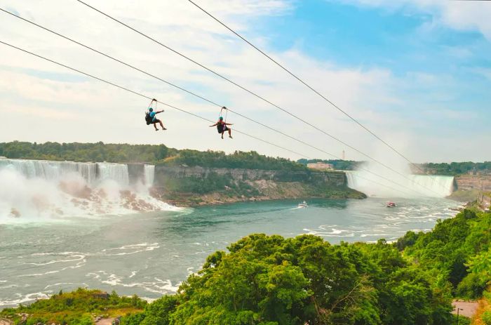 Two zipliners glide down a cable towards a stunning waterfall