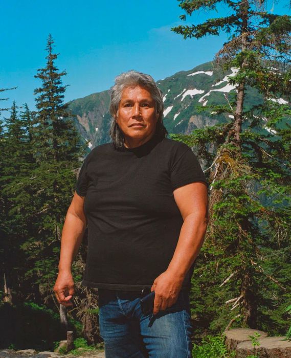 A brown-skinned man with a full head of gray hair stands in front of a grove of trees, dressed in a black T-shirt against a bright blue sky.