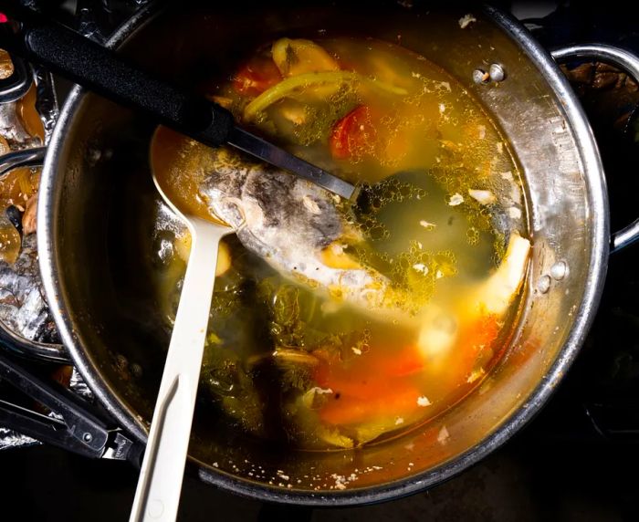 An aerial view of a steel pot featuring two spoons and floating fish heads in a vibrant yellow-orange broth.