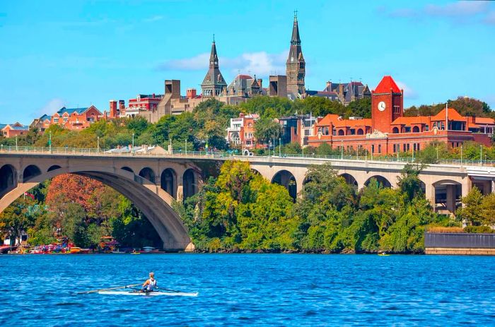 A kayaker approaches a bridge while a large Gothic-style building towers over the riverbank.