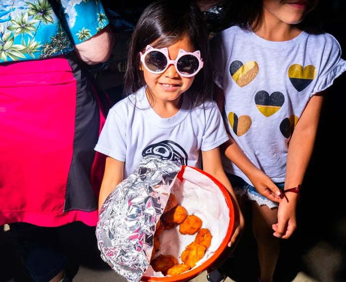 A young girl in pink cat-shaped sunglasses extends a bowl partially filled with golden fry bread lumps.