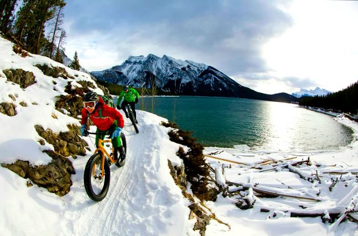 A couple rides fat-tired bikes along a snowy path by a lake in a mountainous setting