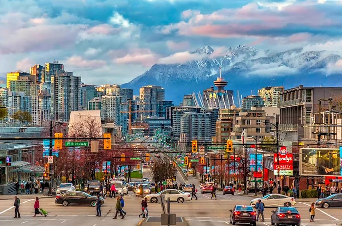 A bustling intersection in Downtown Vancouver where cars pause at traffic lights while pedestrians cross, framed by snow-capped mountains across the water.