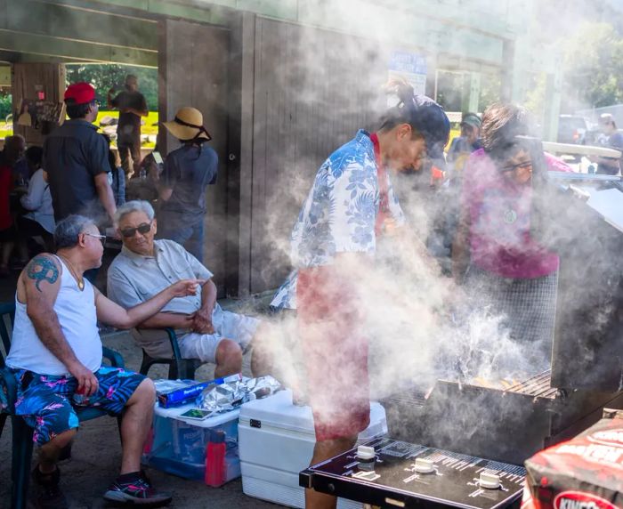 Smoke rises from a grill, where a young man in a baseball cap tends to the food. Nearby, two elderly men sit in lawn chairs, engaged in conversation.