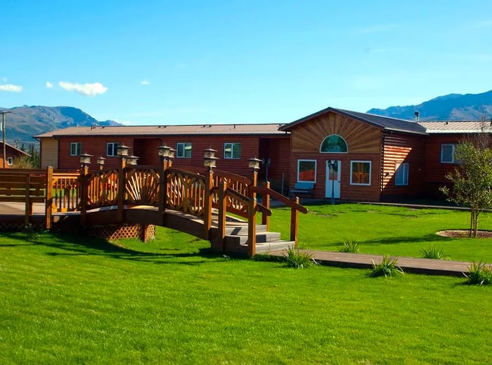 A graceful wooden bridge in front of a rustic building, set against a clear blue sky and lush green grass.