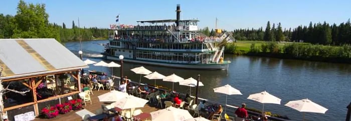 white umbrellas on a deck overlooking a river where a riverboat glides by.