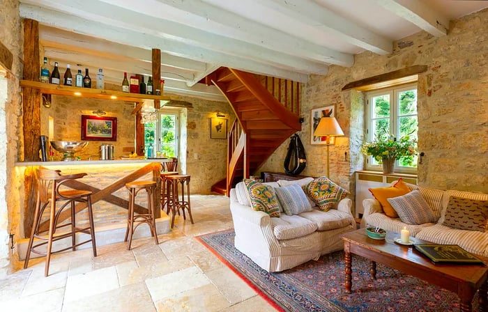 A cozy lounge featuring stone walls, comfortable sofas, and a corner bar. A wooden spiral staircase adds charm in one corner of the room.