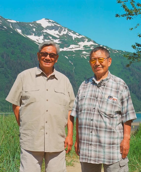 Two elderly men, dressed in beige, stand smiling against a backdrop of a snow-capped mountain surrounded by lush greenery.