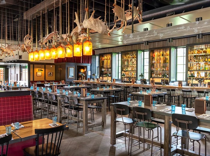 Moose antlers adorn the ceiling of a sleek, modern dining room, featuring long tables that create an industrial vibe.