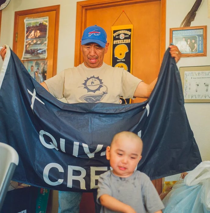 A man in a blue cap meticulously folds a blue flag adorned with white letters, while a small child playfully runs in the foreground.