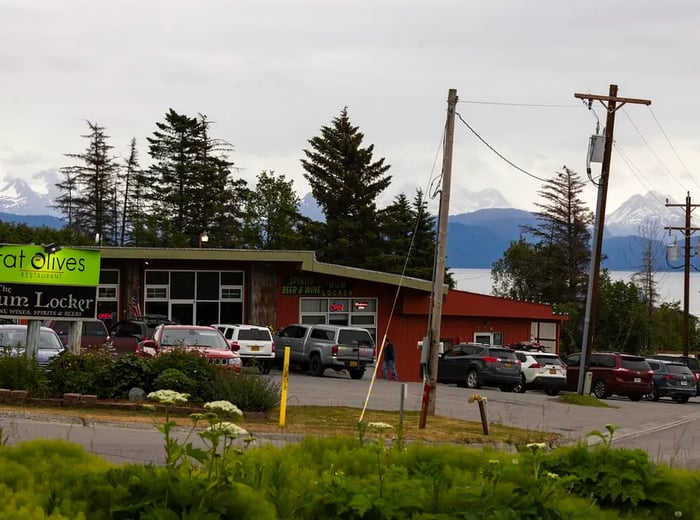 Vehicles are parked outside a quaint red one-story building, framed by snowy mountains and a serene lake in the background.