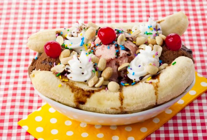 A vibrant banana split featuring three scoops topped with cherries and sprinkles, elegantly arranged on a yellow napkin over a red checkered tablecloth.