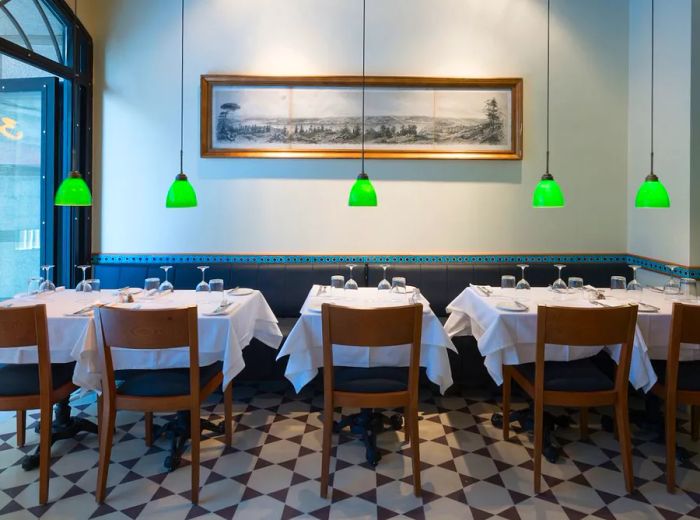 A minimalist dining area featuring tables against a banquette, pendant lights with small green shades, and a long mural adorning the far wall.