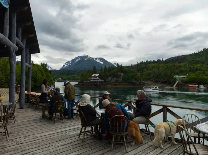 A small group of people and their dogs relax on a wooden deck that overlooks a stunning Alaskan bay.