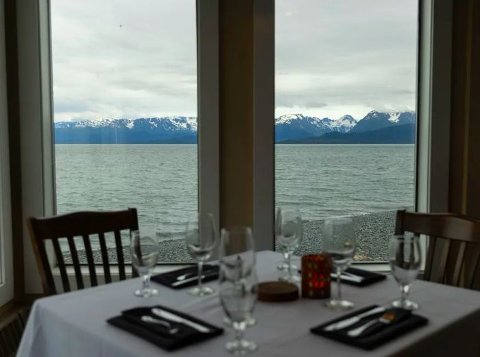 A beautifully set table with a white tablecloth and wine glasses offers a stunning view of a lake and mountains through a picture window.