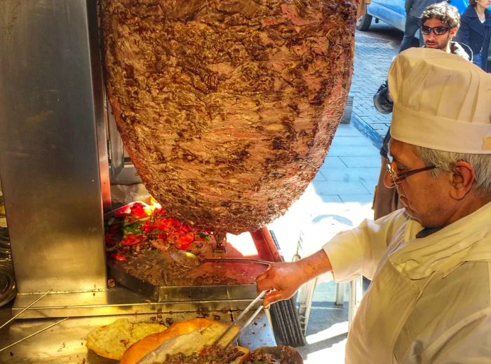 A chef fills fluffy pide with sliced meat, alongside a large rotating spit of meat.