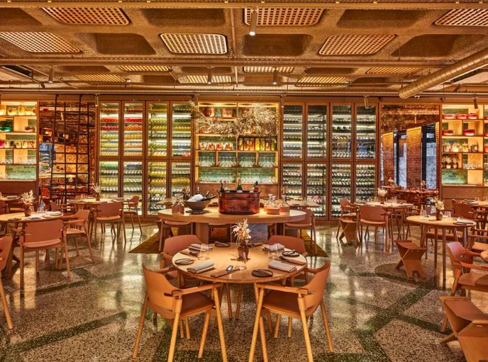 The interior of the restaurant boasts a glamorous design, featuring a wall of wine fridges in the back, ornate mobiles suspended above wooden tables set for dining, and gleaming geometric flooring.