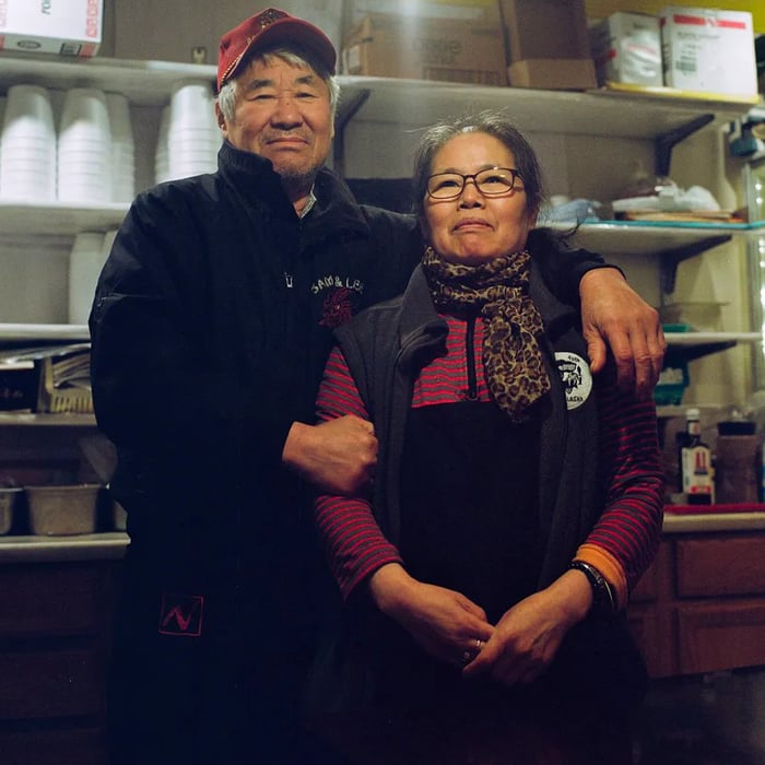 An older Korean man in a red cap stands smiling with his arms around an older Korean woman in front of commercial kitchen shelves.